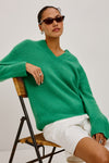 A woman wearing an oversized bright green V-neck knit sweater and white shorts, seated on a woven chair against a plain light gray studio background.