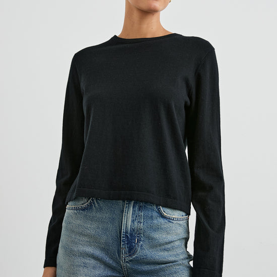 A woman modeling a black long-sleeve crewneck knit top paired with blue jeans, photographed against a light gray studio background.