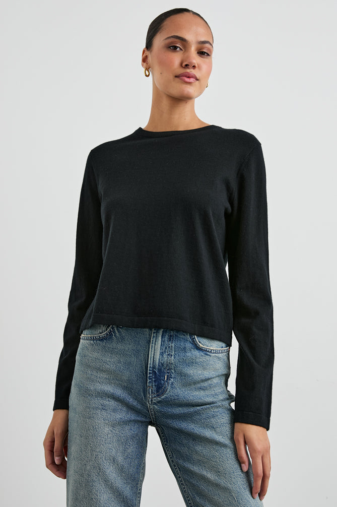 A woman modeling a black long-sleeve crewneck knit top paired with blue jeans, photographed against a light gray studio background.