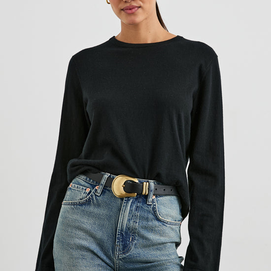 A woman modeling a black long-sleeve crewneck top tucked into light-wash blue jeans with a black belt featuring a large gold buckle, against a light gray studio background.