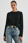 A woman modeling a black long-sleeve crewneck top tucked into light-wash blue jeans with a black belt featuring a large gold buckle, against a light gray studio background.