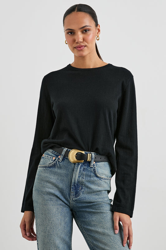 A woman modeling a black long-sleeve crewneck top tucked into light-wash blue jeans with a black belt featuring a large gold buckle, against a light gray studio background.