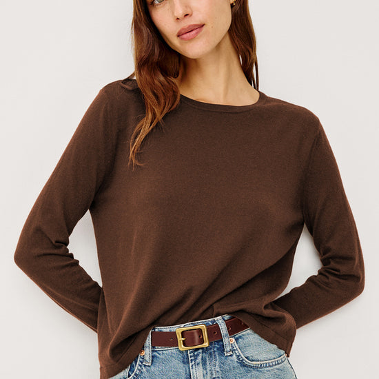 A woman wearing a brown long-sleeve crewneck top and light blue jeans with a brown belt, photographed against a plain light gray studio background.