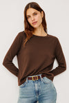 A woman wearing a brown long-sleeve crewneck top and light blue jeans with a brown belt, photographed against a plain light gray studio background.