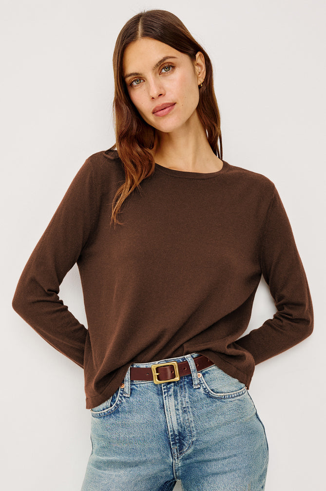 A woman wearing a brown long-sleeve crewneck top and light blue jeans with a brown belt, photographed against a plain light gray studio background.