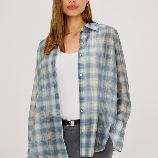 A woman modeling a light blue and white plaid button-up shirt over a white tank top and gray trousers, photographed against a plain light gray studio background.