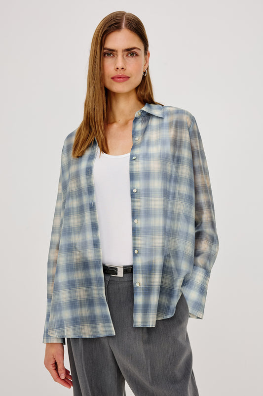 A woman modeling a light blue and white plaid button-up shirt over a white tank top and gray trousers, photographed against a plain light gray studio background.
