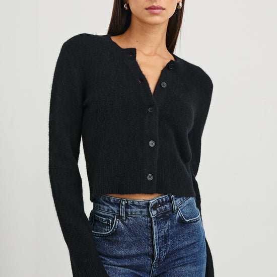 A woman modeling a black long-sleeve cropped button-front cardigan with a split neckline, paired with blue jeans, against a plain light gray studio background.