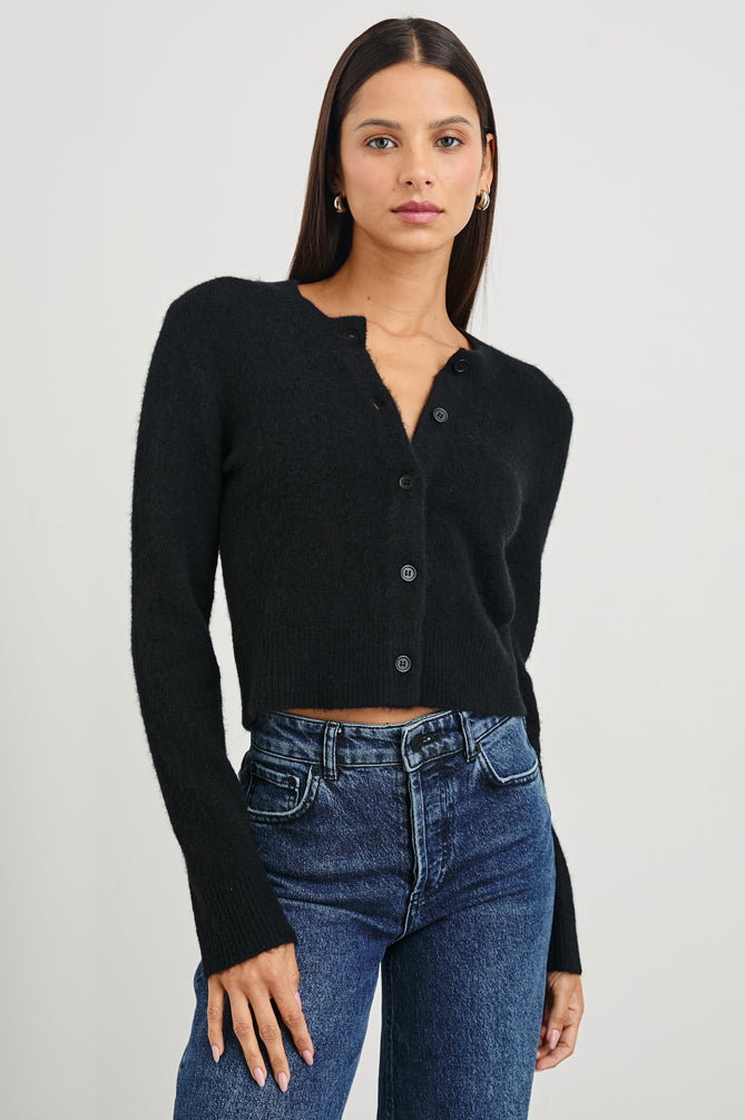 A woman modeling a black long-sleeve cropped button-front cardigan with a split neckline, paired with blue jeans, against a plain light gray studio background.