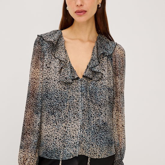A woman modeling a sheer long-sleeve blouse with a ruffled V-neckline and a black, gray, and beige leopard print, paired with black pants against a light gray studio background.