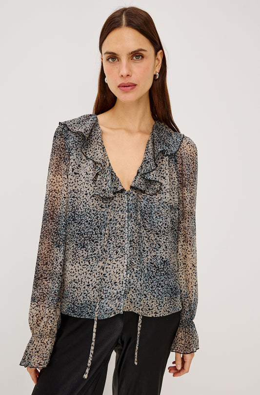 A woman modeling a sheer long-sleeve blouse with a ruffled V-neckline and a black, gray, and beige leopard print, paired with black pants against a light gray studio background.