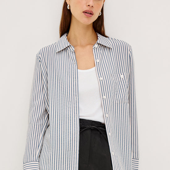 A woman models a long-sleeve button-up shirt with thin vertical navy and white stripes, worn open over a white tank top and black trousers, against a plain light gray studio background.