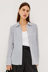 A woman models a long-sleeve button-up shirt with thin vertical navy and white stripes, worn open over a white tank top and black trousers, against a plain light gray studio background.