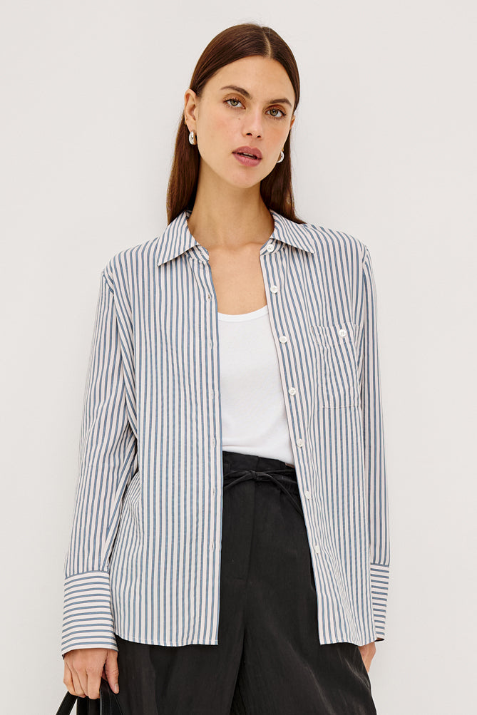 A woman models a long-sleeve button-up shirt with thin vertical navy and white stripes, worn open over a white tank top and black trousers, against a plain light gray studio background.