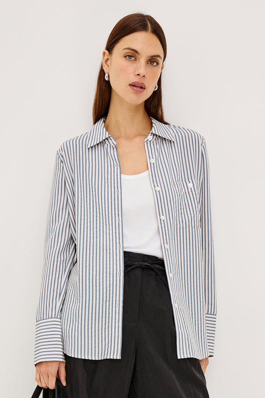 A woman models a long-sleeve button-up shirt with thin vertical navy and white stripes, worn open over a white tank top and black trousers, against a plain light gray studio background.
