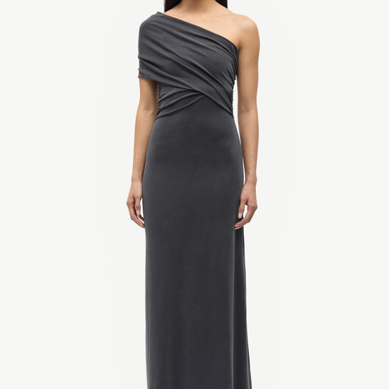A model wearing a dark gray, one-shoulder draped maxi dress with a fitted waist and flowing skirt, paired with black pointed-toe heels, against a plain light gray background.