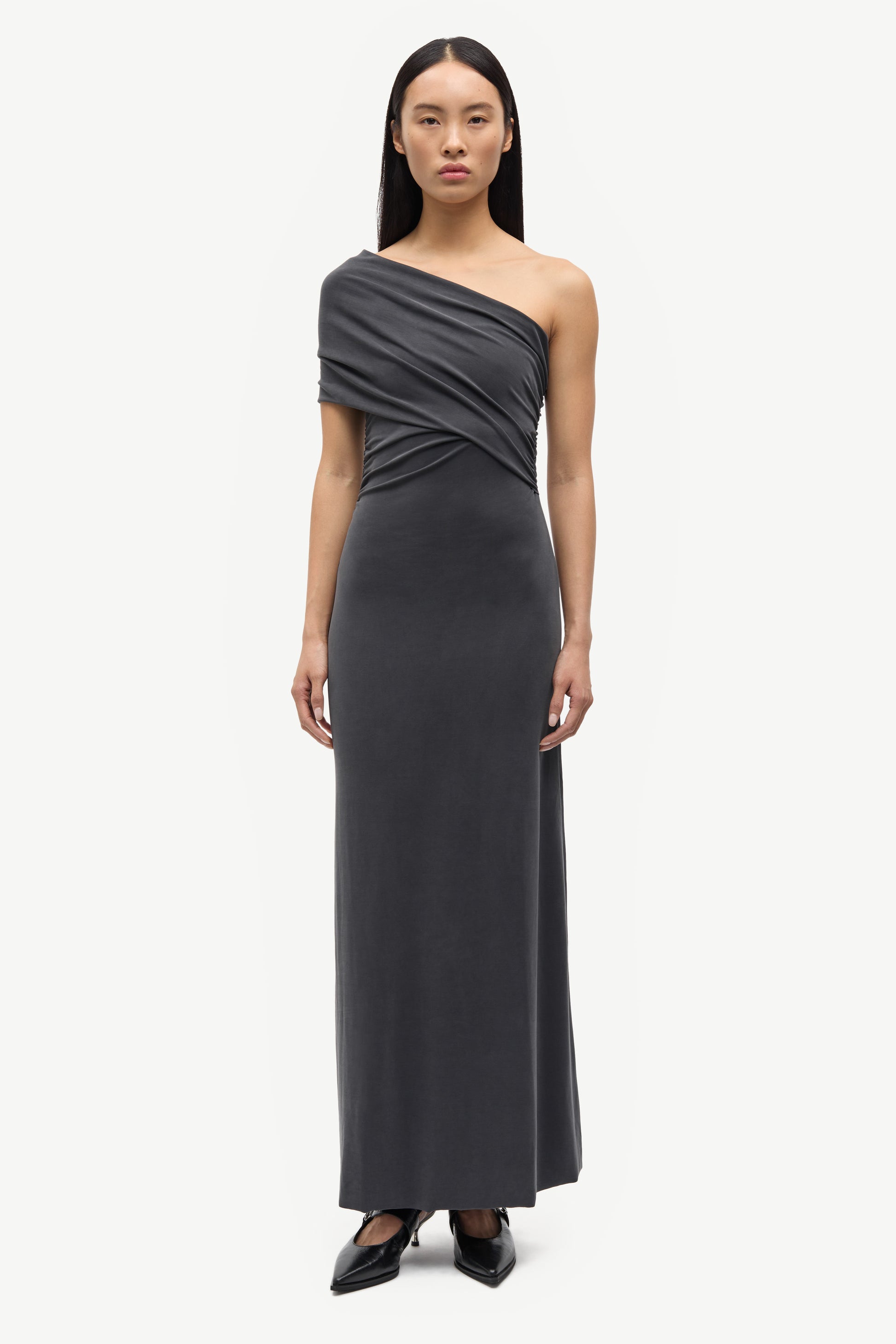 A model wearing a dark gray, one-shoulder draped maxi dress with a fitted waist and flowing skirt, paired with black pointed-toe heels, against a plain light gray background.