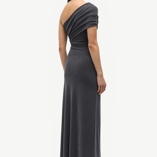 A woman shown from the back wearing a dark charcoal gray one-shoulder maxi dress with a draped neckline and short sleeves, paired with black pointed-toe heels, against a light gray studio background.