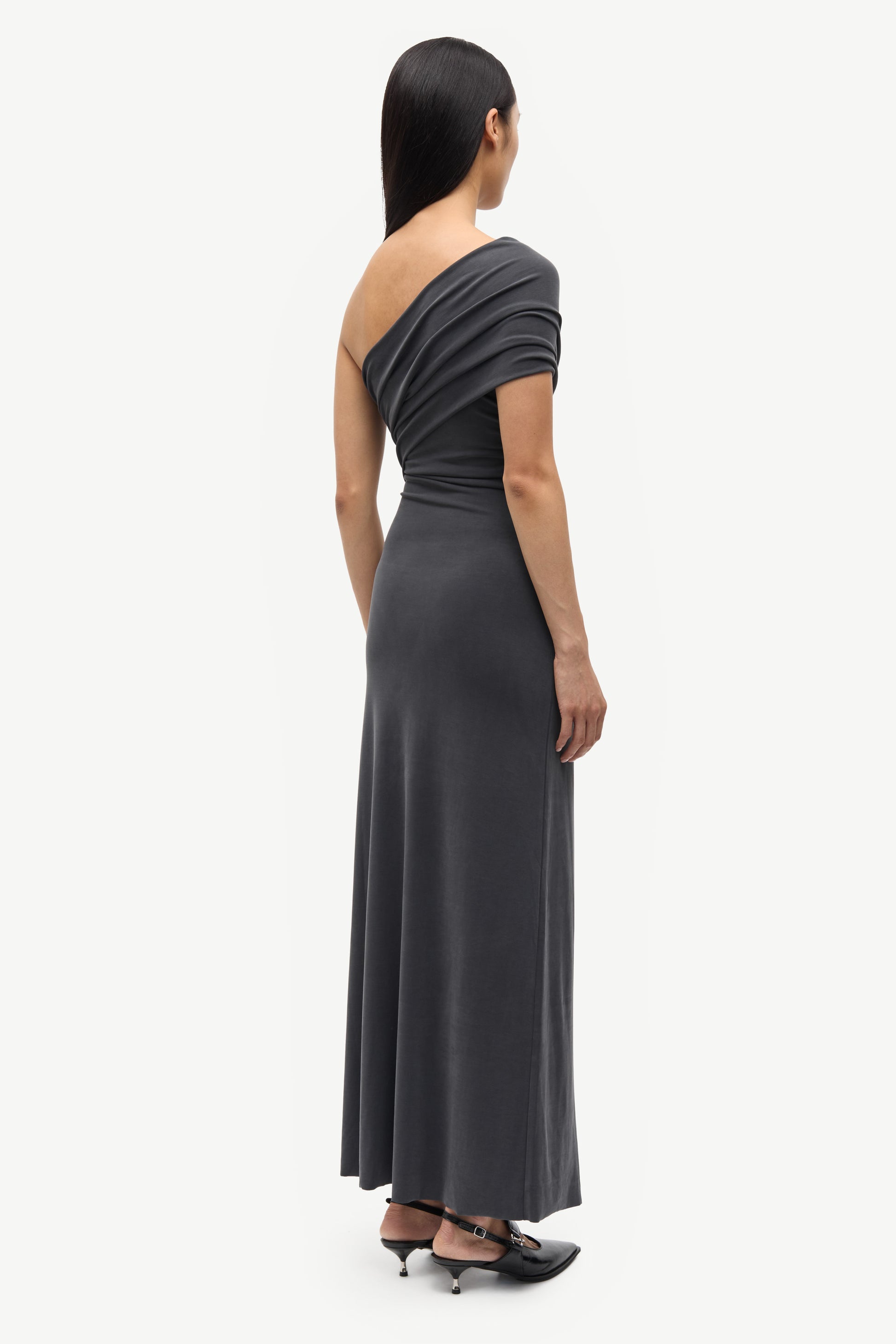 A woman shown from the back wearing a dark charcoal gray one-shoulder maxi dress with a draped neckline and short sleeves, paired with black pointed-toe heels, against a light gray studio background.