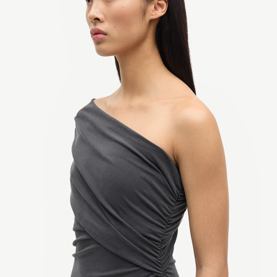 A woman wearing a dark gray one-shoulder ruched dress, shown in a side profile against a plain light gray studio background.