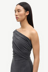 A woman wearing a dark gray one-shoulder ruched dress, shown in a side profile against a plain light gray studio background.
