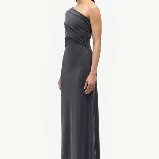 A woman modeling a dark gray one-shoulder maxi dress with a draped bodice, paired with black pointed-toe flats, against a plain light gray studio background.