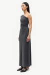 A woman modeling a dark gray one-shoulder maxi dress with a draped bodice, paired with black pointed-toe flats, against a plain light gray studio background.