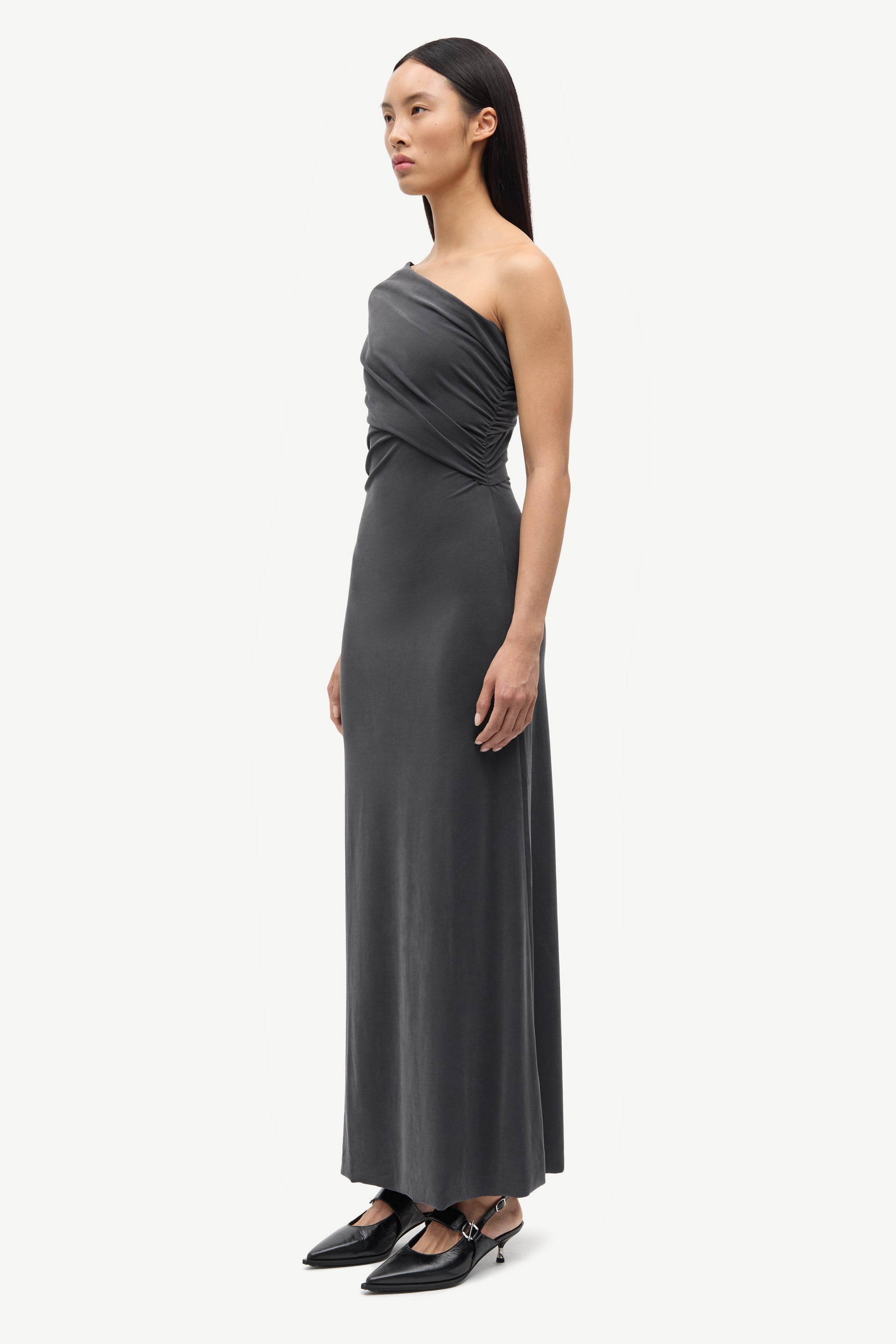 A woman modeling a dark gray one-shoulder maxi dress with a draped bodice, paired with black pointed-toe flats, against a plain light gray studio background.