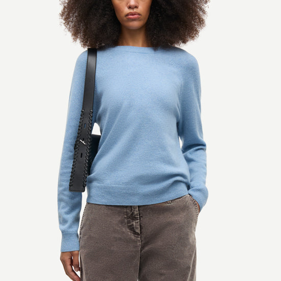 A model with dark curly hair wearing a light blue knit sweater, brown corduroy trousers, and carrying a black shoulder bag, photographed against a solid off-white background.