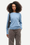 A model with dark curly hair wearing a light blue knit sweater, brown corduroy trousers, and carrying a black shoulder bag, photographed against a solid off-white background.