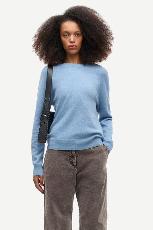 A model with dark curly hair wearing a light blue knit sweater, brown corduroy trousers, and carrying a black shoulder bag, photographed against a solid off-white background.