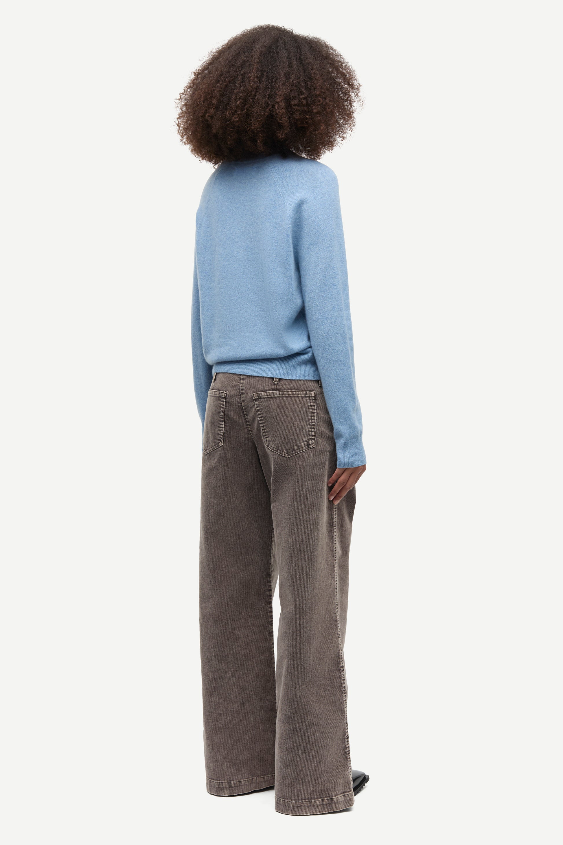 A child model shown from the back wearing a light blue long-sleeve knit top and wide-leg brown corduroy trousers, set against a plain light gray studio background.