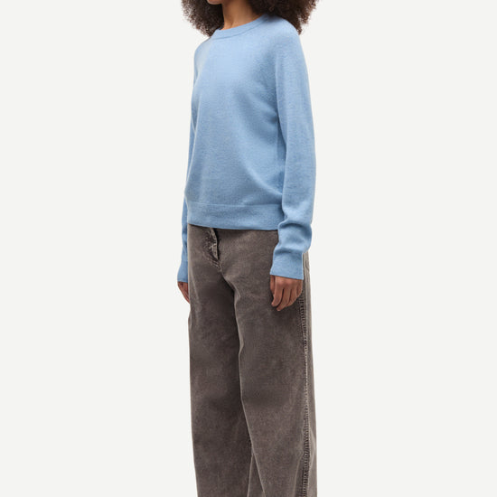 A model wearing a light blue crewneck sweater and wide-leg brown corduroy trousers, standing in profile against a plain light gray background.