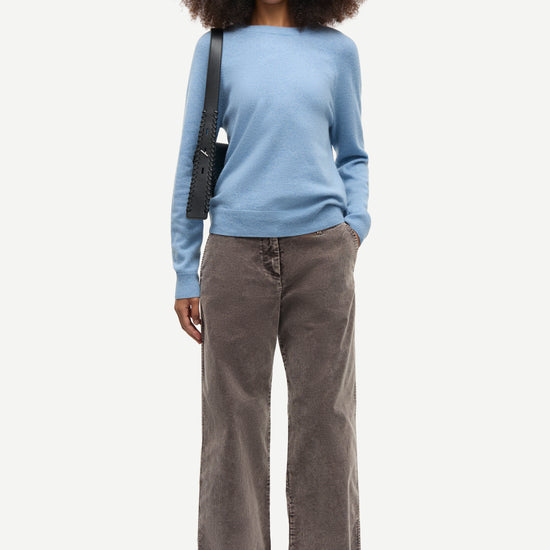 A model with curly dark hair stands against a light gray studio background wearing a light blue long-sleeve knit top, wide-leg brown corduroy pants, and black shoes, with a black shoulder bag.