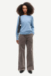 A model with curly dark hair stands against a light gray studio background wearing a light blue long-sleeve knit top, wide-leg brown corduroy pants, and black shoes, with a black shoulder bag.