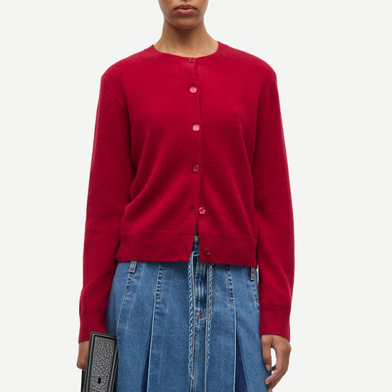 A model wearing a vibrant red button-up cardigan and a blue denim skirt, holding a black textured handbag against a plain light gray background.