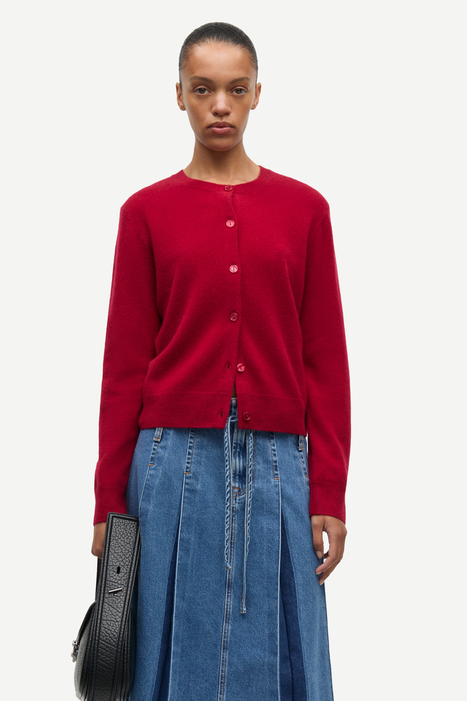 A model wearing a vibrant red button-up cardigan and a blue denim skirt, holding a black textured handbag against a plain light gray background.