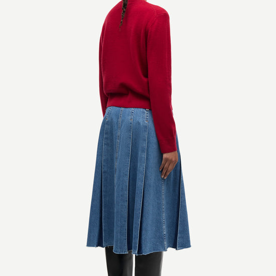 A model shown from the back wearing a red knit sweater, a blue pleated denim skirt, and black leather boots against a light gray studio background.