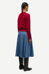 A model shown from the back wearing a red knit sweater, a blue pleated denim skirt, and black leather boots against a light gray studio background.