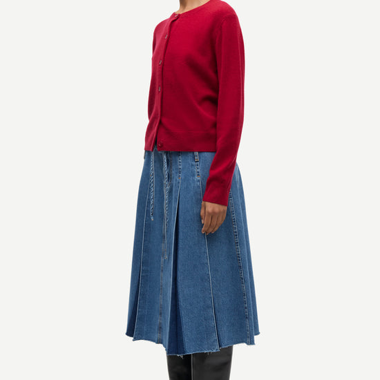 A model is shown in profile wearing a dark red long-sleeve knit cardigan, a blue denim pleated midi skirt, and black leather boots against a light gray studio background.
