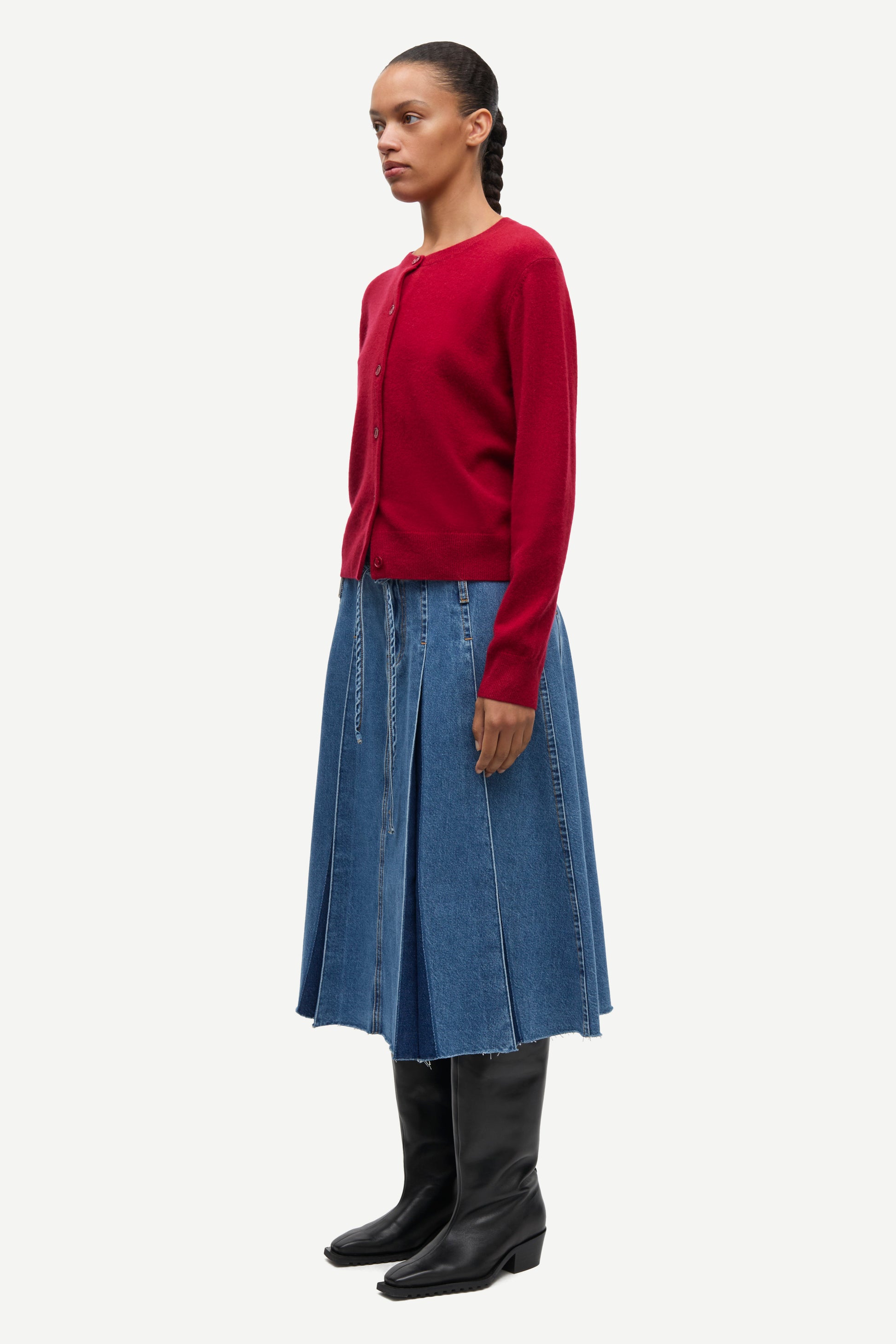 A model is shown in profile wearing a dark red long-sleeve knit cardigan, a blue denim pleated midi skirt, and black leather boots against a light gray studio background.