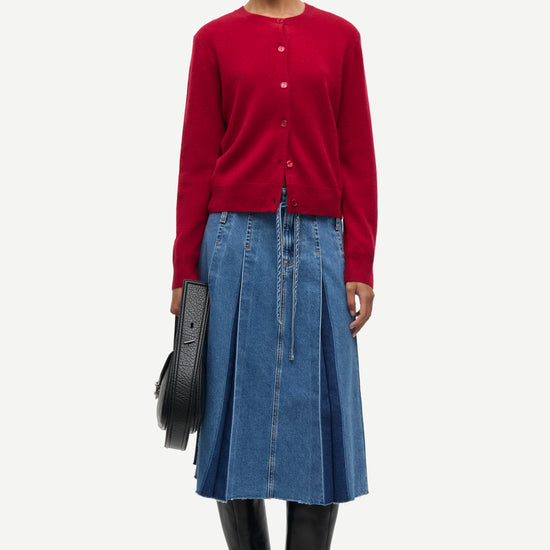 A model stands against a light gray studio background wearing a red button-front cardigan, a blue pleated denim skirt, black knee-high boots, and holding a black circular clutch.