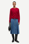 A model stands against a light gray studio background wearing a red button-front cardigan, a blue pleated denim skirt, black knee-high boots, and holding a black circular clutch.