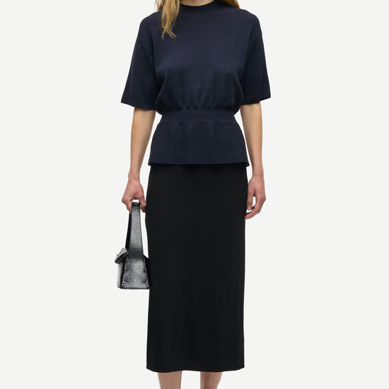 A model stands against a light gray studio background wearing a dark navy short-sleeve peplum top and a matching dark midi skirt, holding a small dark handbag and wearing olive-green and black sneakers.