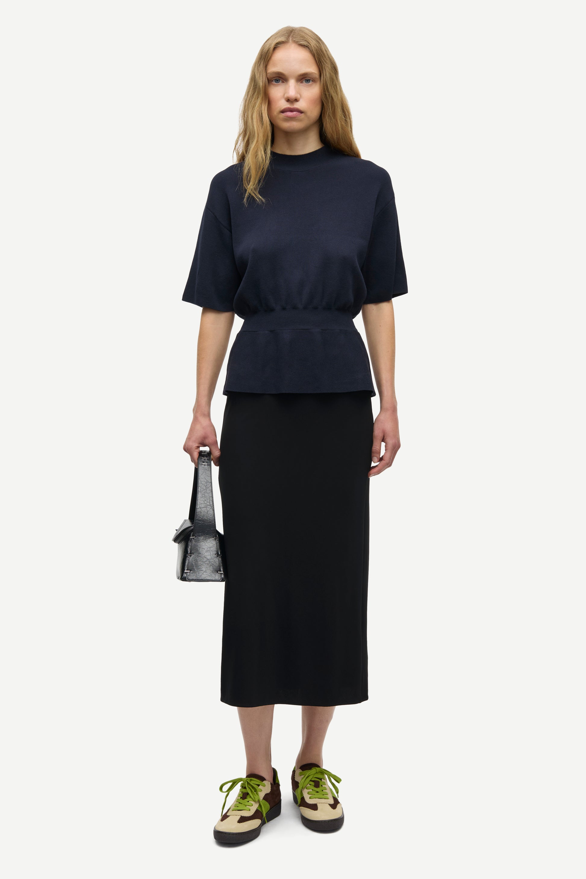 A model stands against a light gray studio background wearing a dark navy short-sleeve peplum top and a matching dark midi skirt, holding a small dark handbag and wearing olive-green and black sneakers.