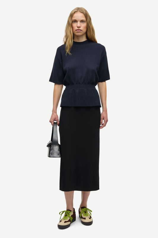 A model stands against a light gray studio background wearing a dark navy short-sleeve peplum top and a matching dark midi skirt, holding a small dark handbag and wearing olive-green and black sneakers.