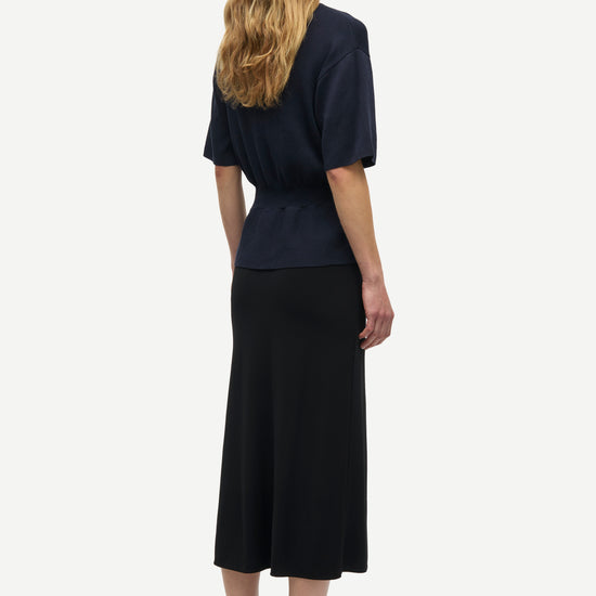 A woman shown from the back wearing a dark navy short-sleeve top and a long black midi skirt, paired with olive green sandals, against a plain light gray background.