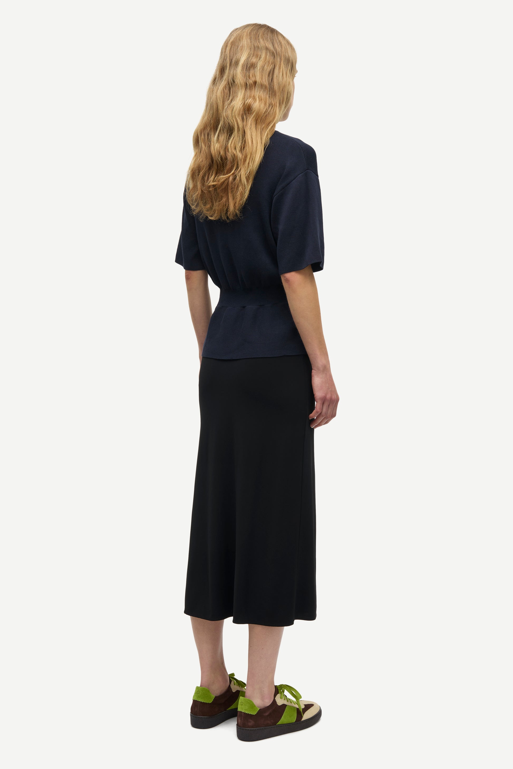 A woman shown from the back wearing a dark navy short-sleeve top and a long black midi skirt, paired with olive green sandals, against a plain light gray background.