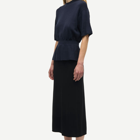 A model is shown from the side wearing a dark navy blue short-sleeve top with a peplum waist, a long black skirt, and olive green sneakers against a plain light gray background.