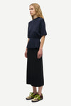 A model is shown from the side wearing a dark navy blue short-sleeve top with a peplum waist, a long black skirt, and olive green sneakers against a plain light gray background.